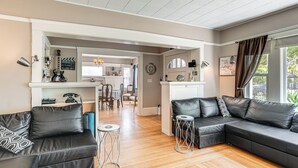 A modern living room with a black leather sofa, a dining area with a wooden table, and a kitchen with a white countertop.