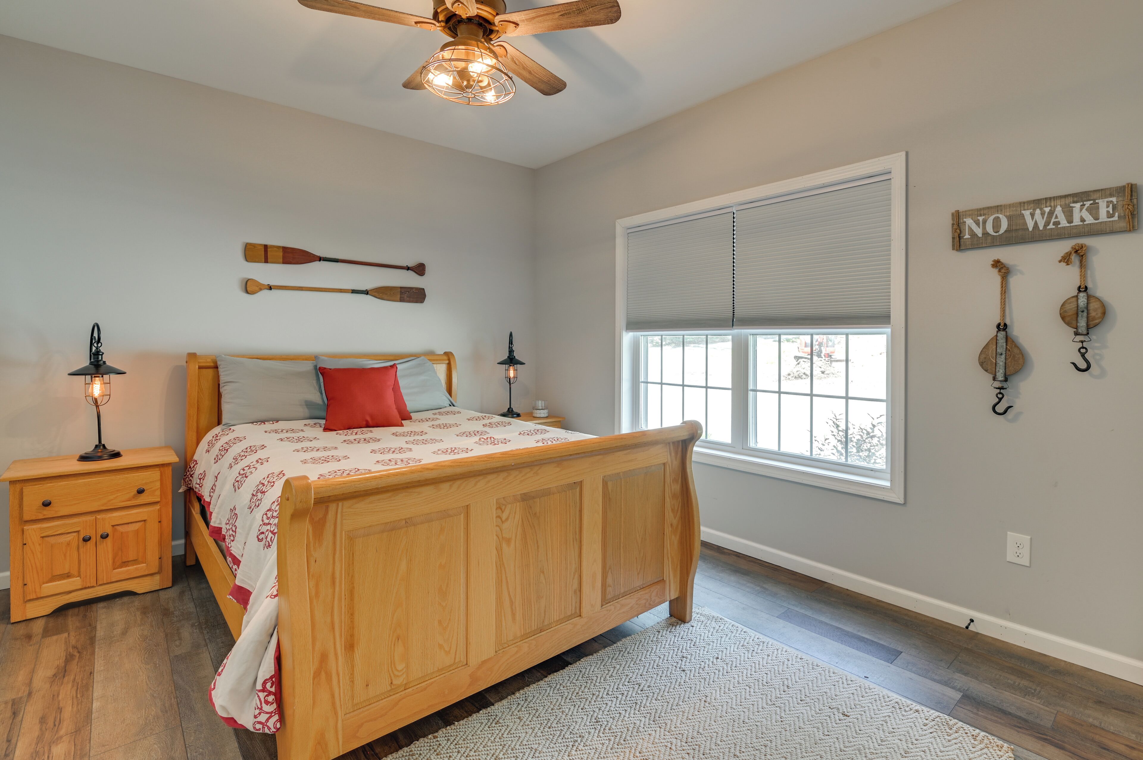 A bedroom with a wooden bed, two nightstands, a ceiling fan, a window with blinds, and wall decorations including paddles and a 'NO WAKE' sign.
