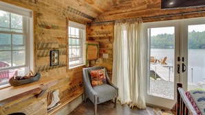 A log cabin room with a wooden ceiling, a large window overlooking a lake, a chair with a cushion, and a small table with a bowl of pinecones. - Cozy Cabin on Crooked Lake: Swim, Kayak & Explore! (Averill Park)