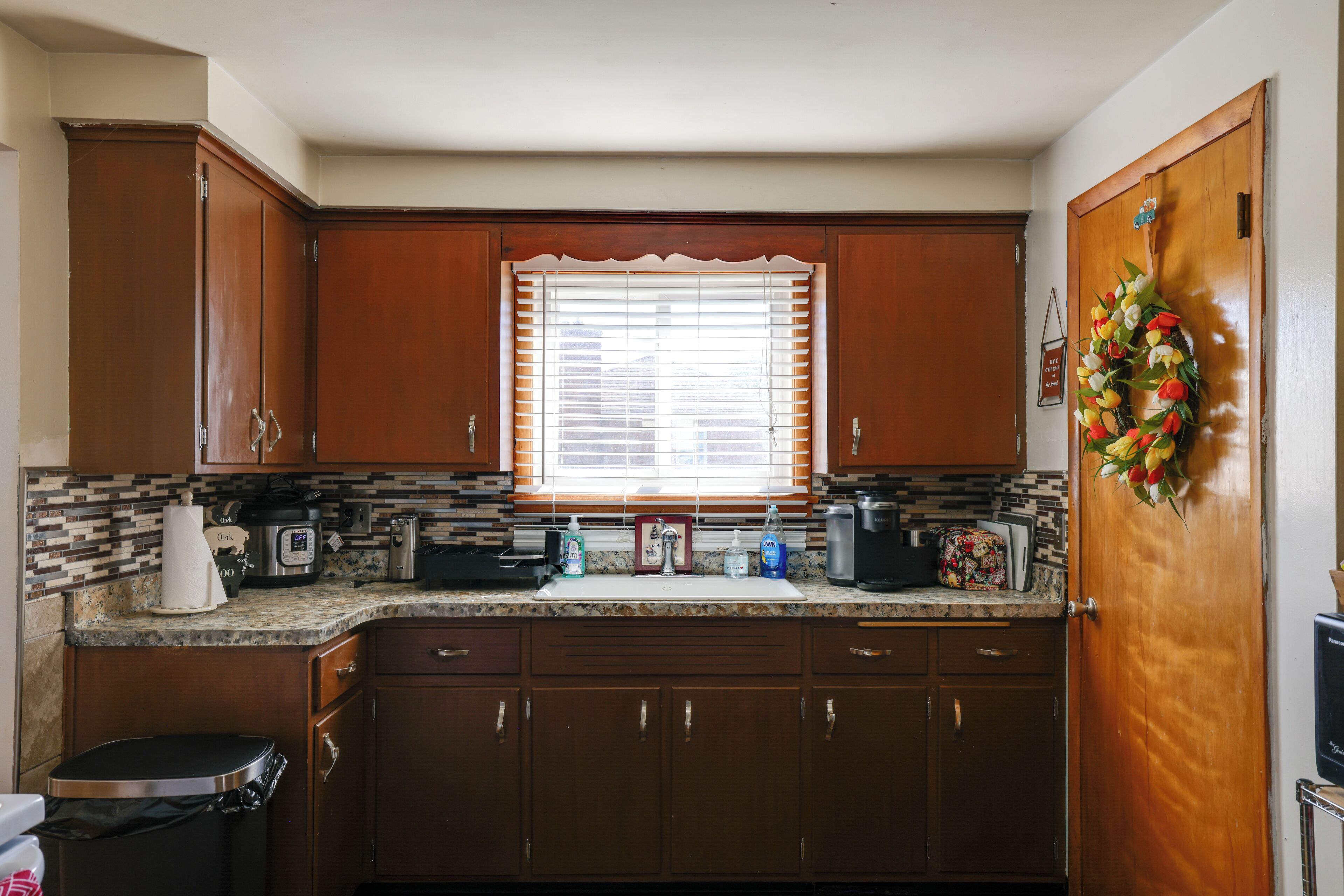 A kitchen with wooden cabinets, a window with blinds, a sink, and a trash can.