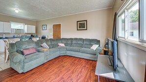 Un salon avec un canapé d’angle, un plancher en bois, un téléviseur à écran plat et une fenêtre avec des rideaux.