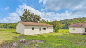 Une maison en bois blanche, d’un seul étage, avec un toit brun, entourée d’une cour verdoyante et bordée, au loin, d’une colline boisée.