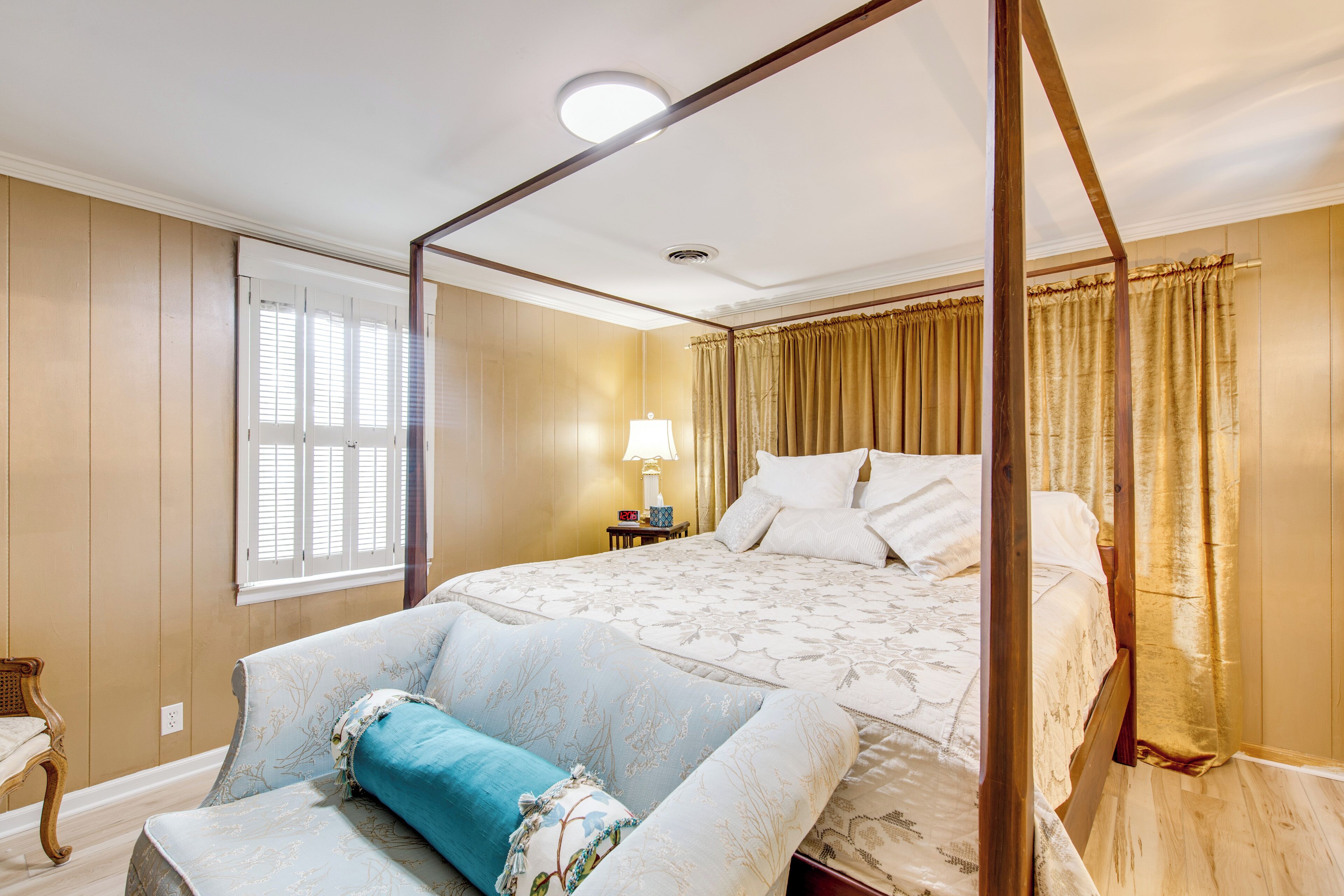 A bedroom with a four-poster bed, a bedside table with a lamp, and a window with blinds.