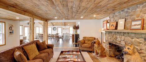 A spacious living room with a wooden ceiling, a stone fireplace, and a dining area in the background.