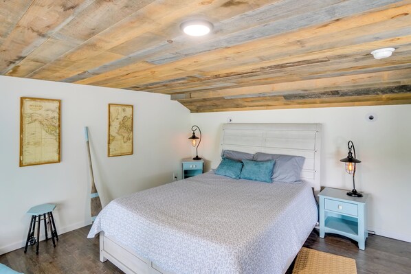 A bedroom with a wooden ceiling, a bed with a white headboard, two nightstands with lamps, and two framed world maps on the wall. - Spectacular Crooked Lake Home: Wake Up to Wonder! (Averill Park)
