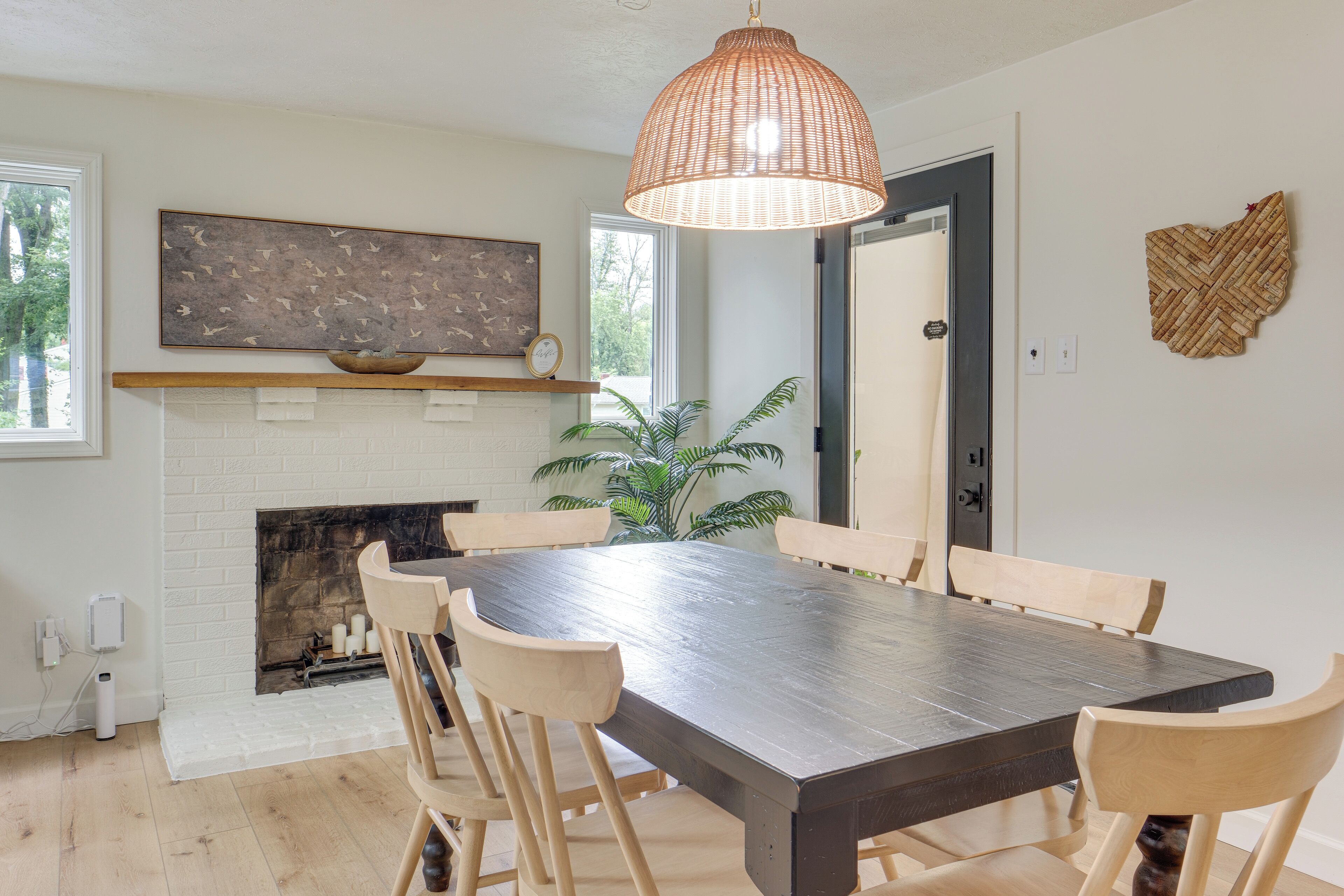 Een eetruimte met een grote houten tafel, zes stoelen, een hanglamp van geweven materiaal, een open haard en een kunstwerk aan de muur.