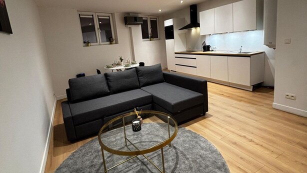 Una sala de estar moderna con un sofá modular, una mesa de centro redonda y una zona de cocina con armarios blancos y una campana extractora negra.