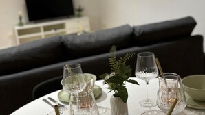 Una mesa de comedor dispuesta con cristalería y una planta en maceta, con un sofá y televisor al fondo.