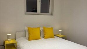 Una cama con ropa blanca y dos almohadas amarillas, flanqueada por dos mesitas de noche con lámparas, en una habitación con ventana sobre la cama.