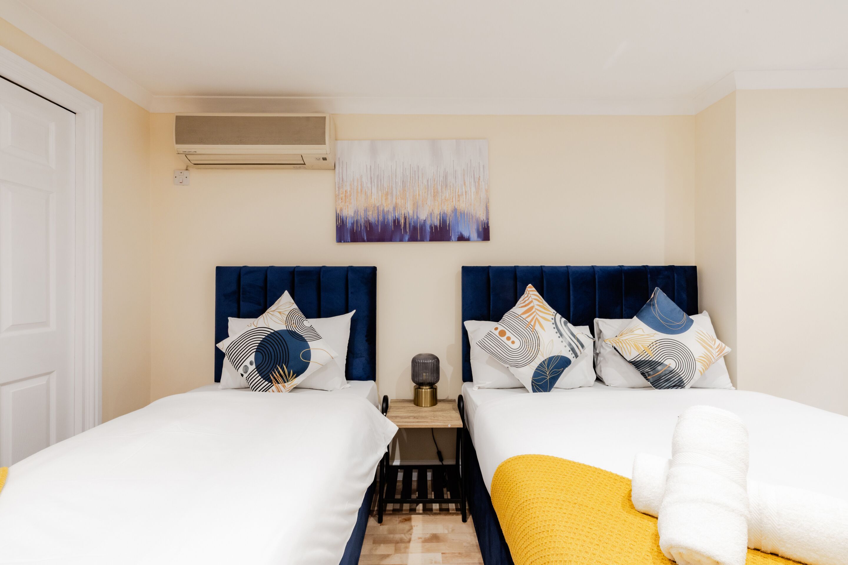 Une chambre d’hôtel avec deux lits, un climatiseur fixé au mur, un tableau abstrait décoratif et une petite table avec une lampe.