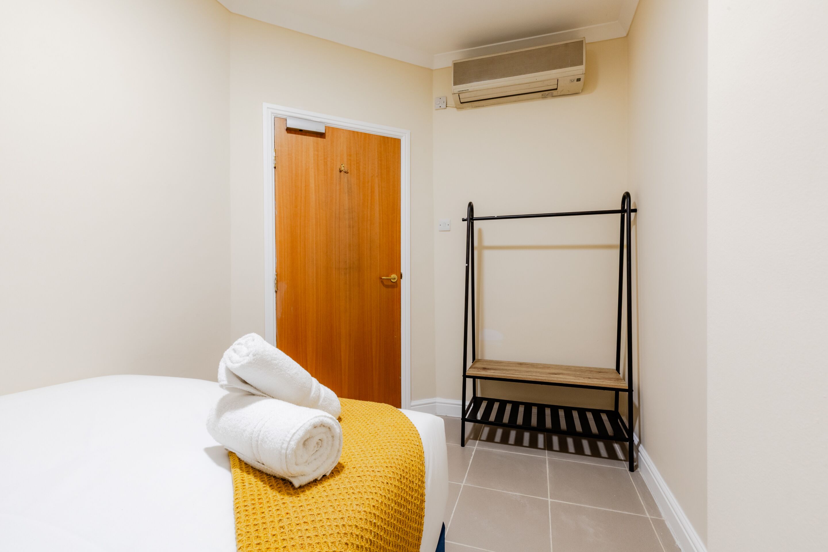 A hotel room with a bed, a wooden door, a black metal drying rack, and a wall-mounted air conditioner.