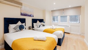 A hotel room with two beds, each with a blue headboard and white bedding, a yellow throw blanket, and a painting on the wall. - Comfy Apartments in Queensway (London)