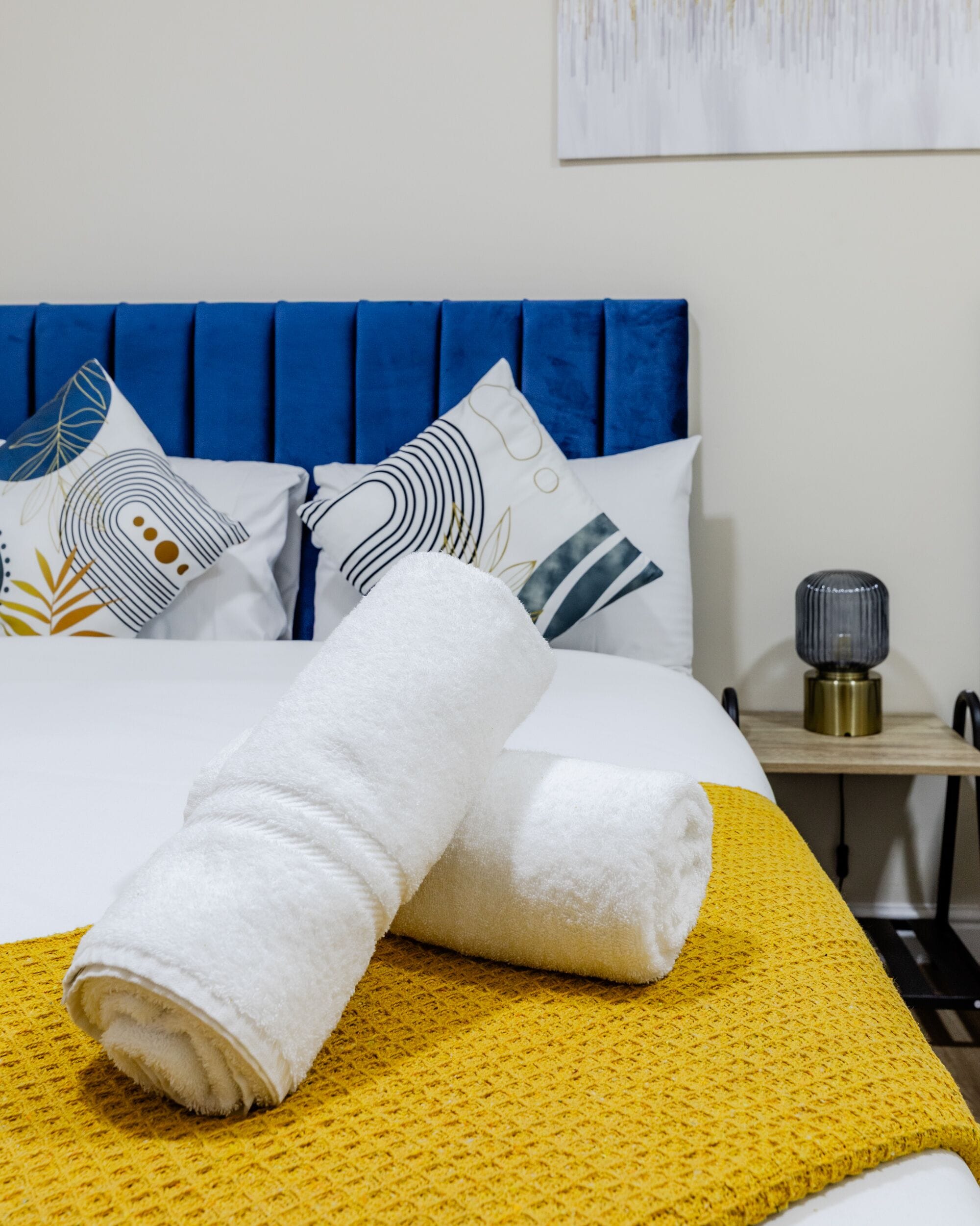 Un lit bien fait, agrémenté d’une serviette blanche roulée, d’un plaid jaune et de coussins décoratifs aux motifs géométriques.