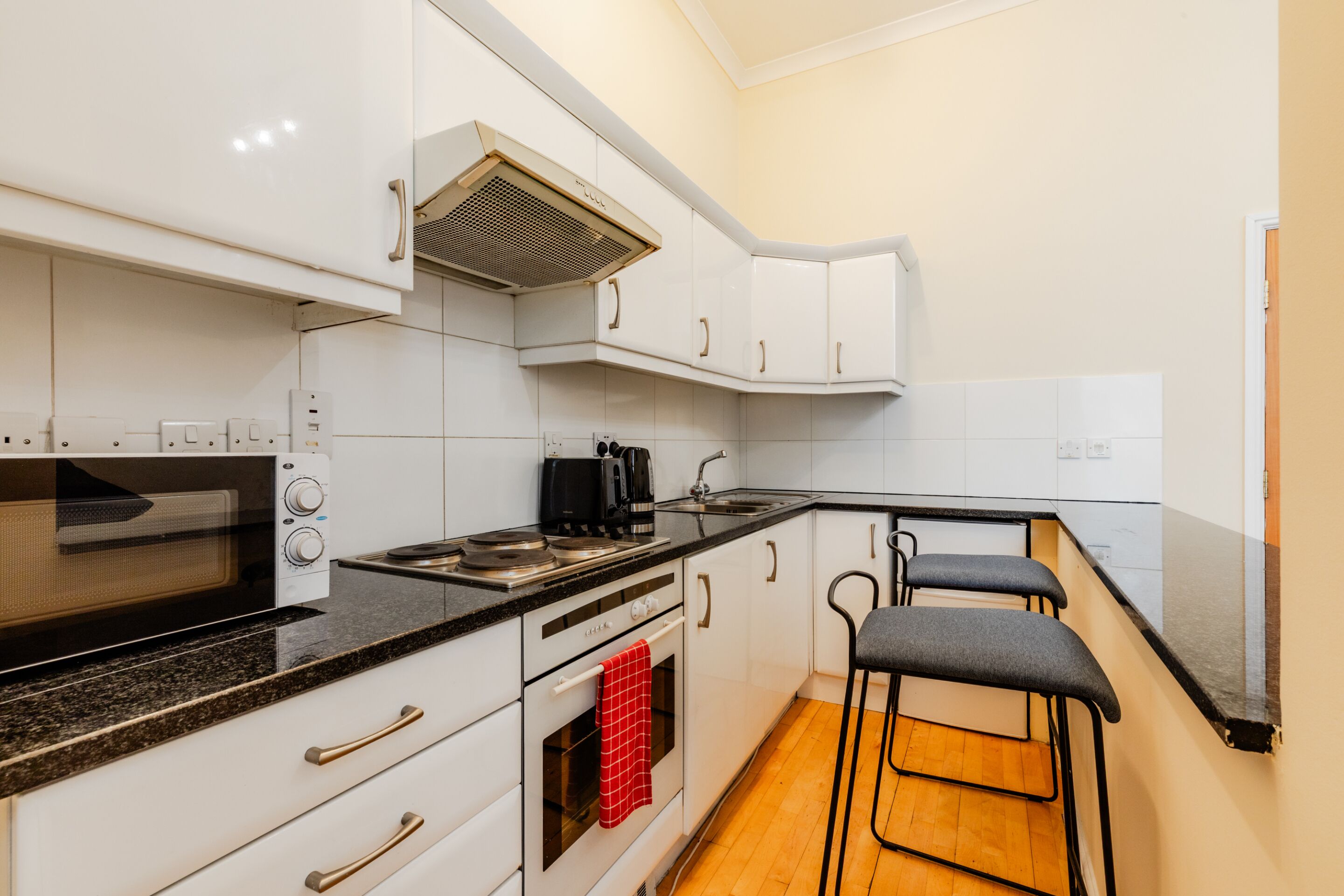 Una cocina compacta con armarios blancos, un microondas, una estufa y un lavamanos. Hay dos taburetes de bar y una toalla roja colgada en la puerta del horno.