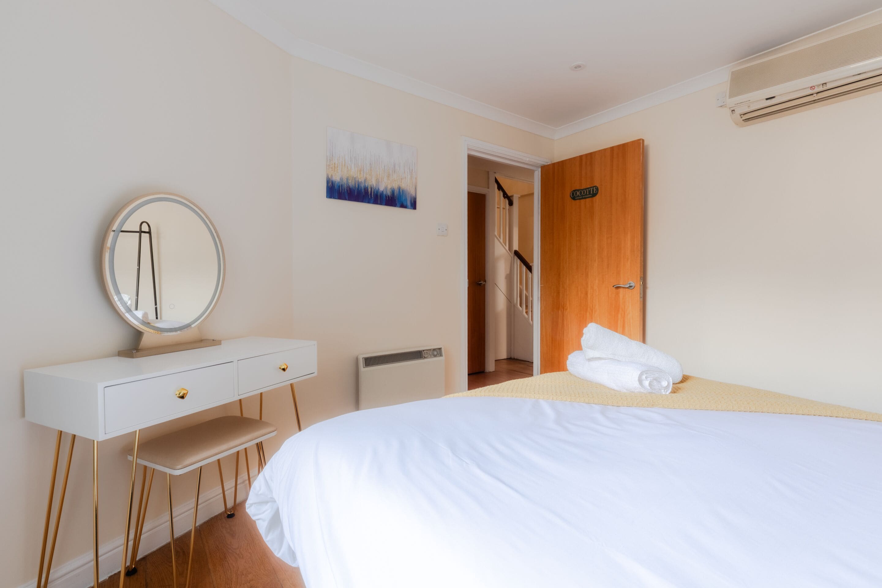 A hotel room with a bed, a bedside table with a mirror, a small bench, a door, and a wall-mounted air conditioner.