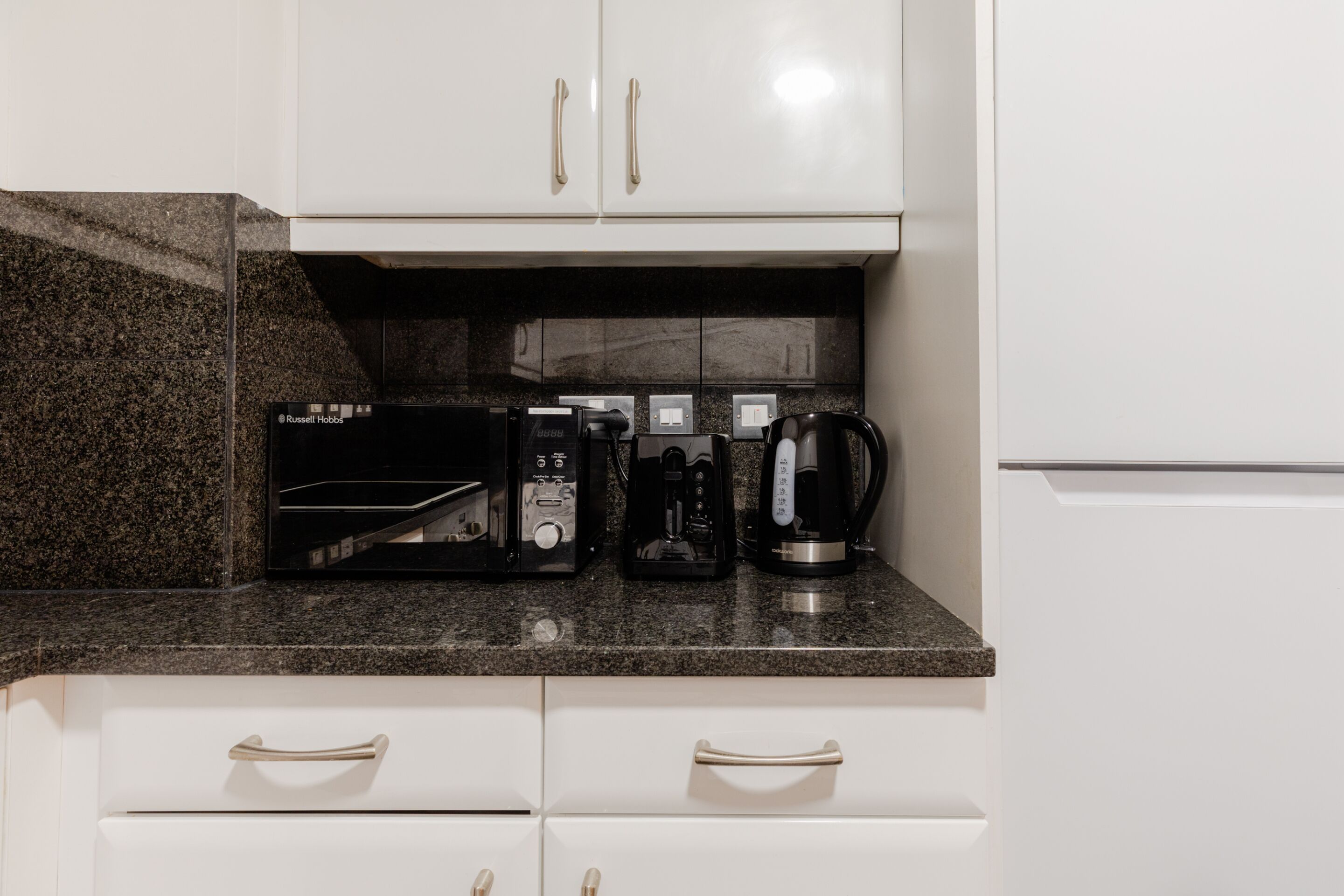 Eine Küchenzeile mit Mikrowelle, Kaffeemaschine und Toaster auf einer Arbeitsplatte aus Granit, darüber befinden sich weiße Schränke mit goldenen Griffen.