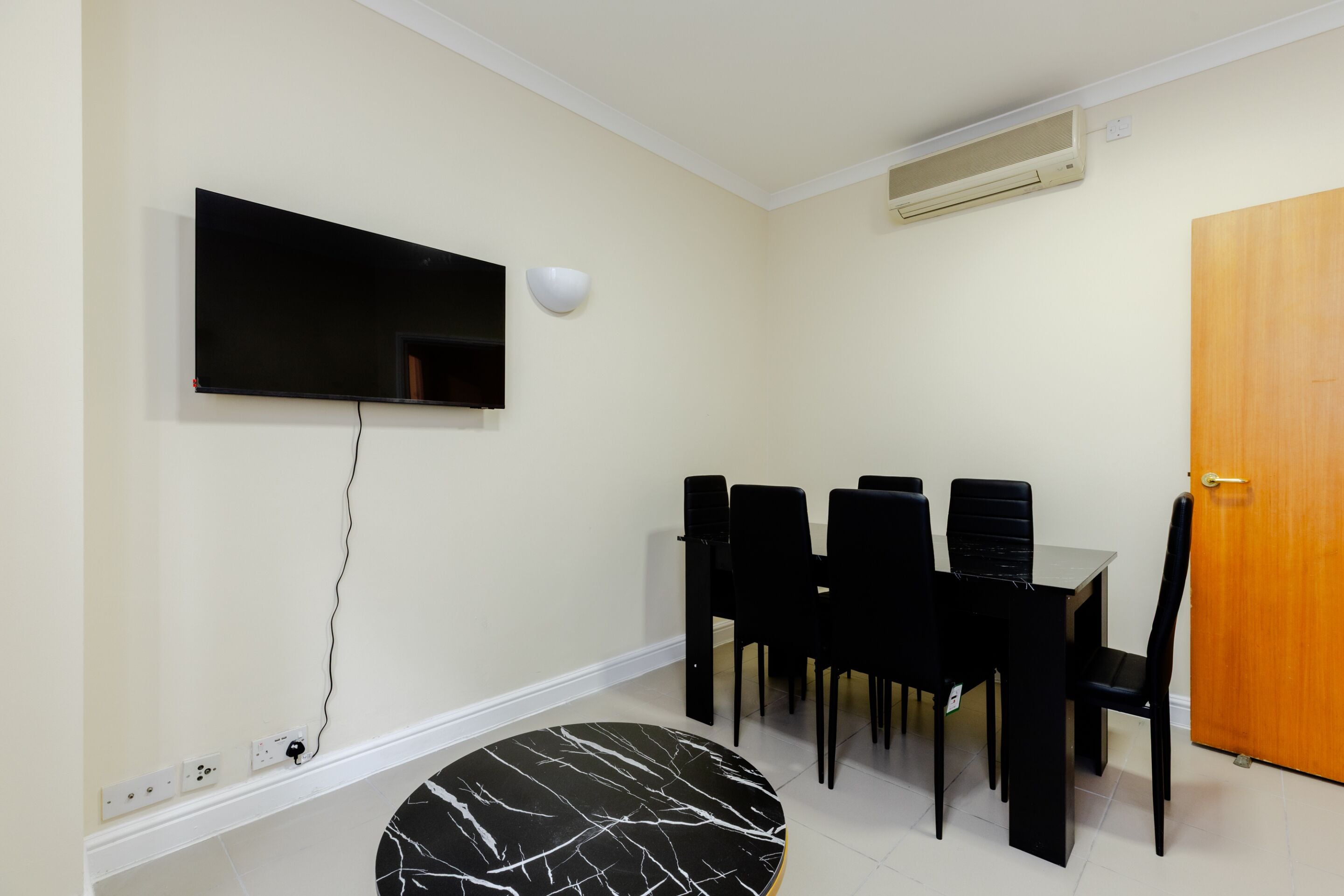 A dining area with a TV mounted on the wall, a round table with chairs, and a wooden door.