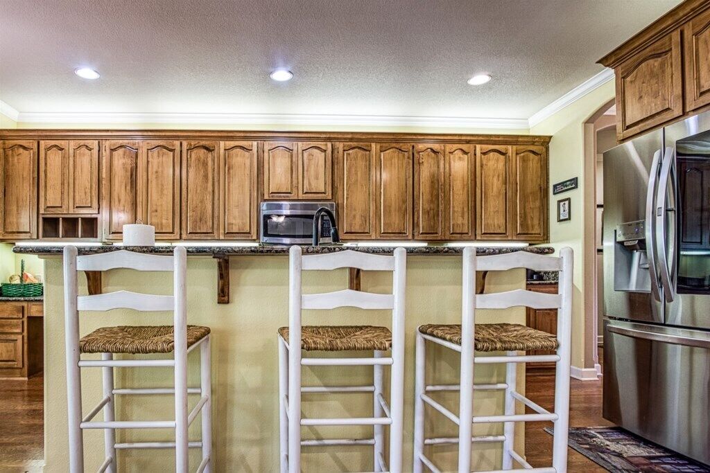 Una cocina con armarios de madera, un refrigerador de acero inoxidable y tres taburetes blancos con asientos de mimbre.