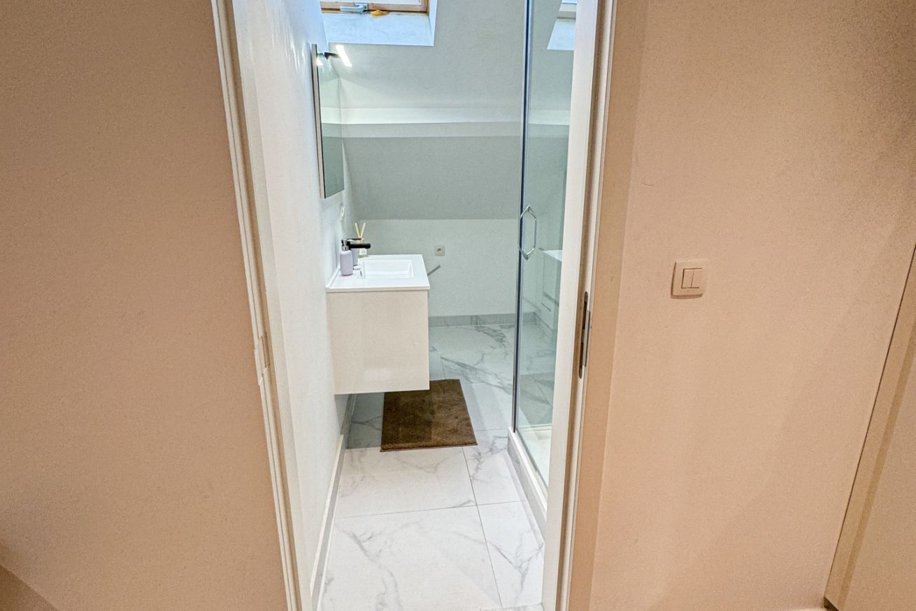A narrow hallway leading to a bathroom with a glass shower door, a white sink, and a marble floor.