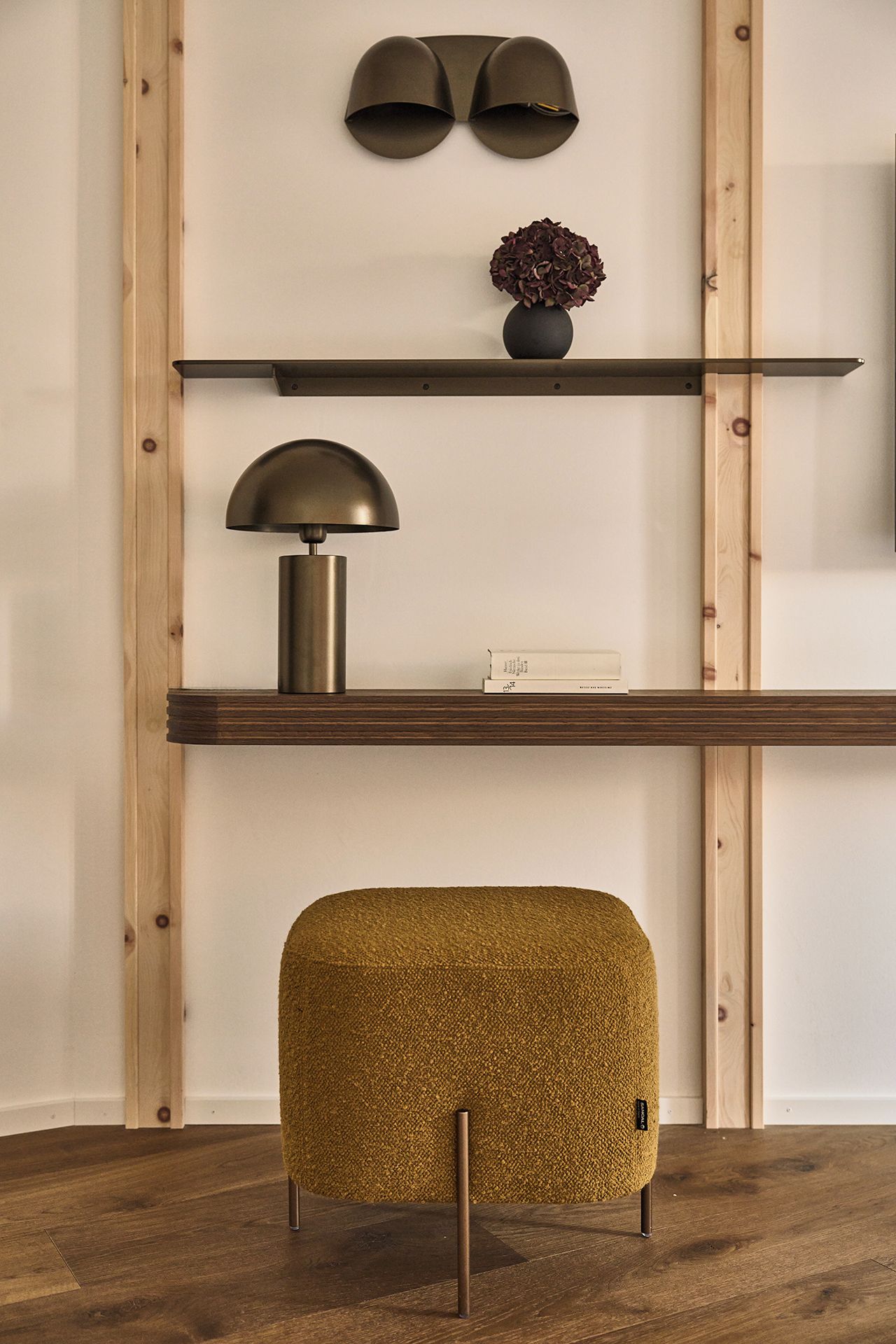 A room with a wooden floor, a shelf with a lamp, a vase with flowers, and a textured ottoman.