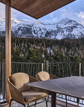 Een overdekt terras met twee stoelen en een tafel, met uitzicht op besneeuwde bergen en een bebost gebied.