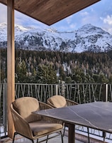 Un espacio exterior techado con dos sillas y una mesa, con vistas a montañas nevadas y una zona boscosa.