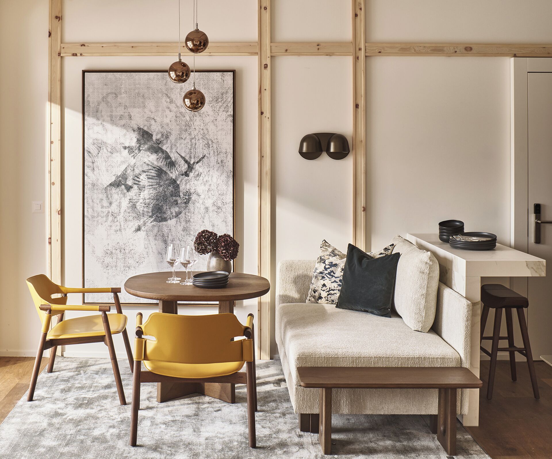 A modern living room with a round wooden table, two yellow chairs, a beige sofa with a dark pillow, and a large abstract painting on the wall.