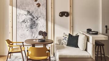A modern living room with a round wooden table, two yellow chairs, a beige sofa with a dark pillow, and a large abstract painting on the wall.