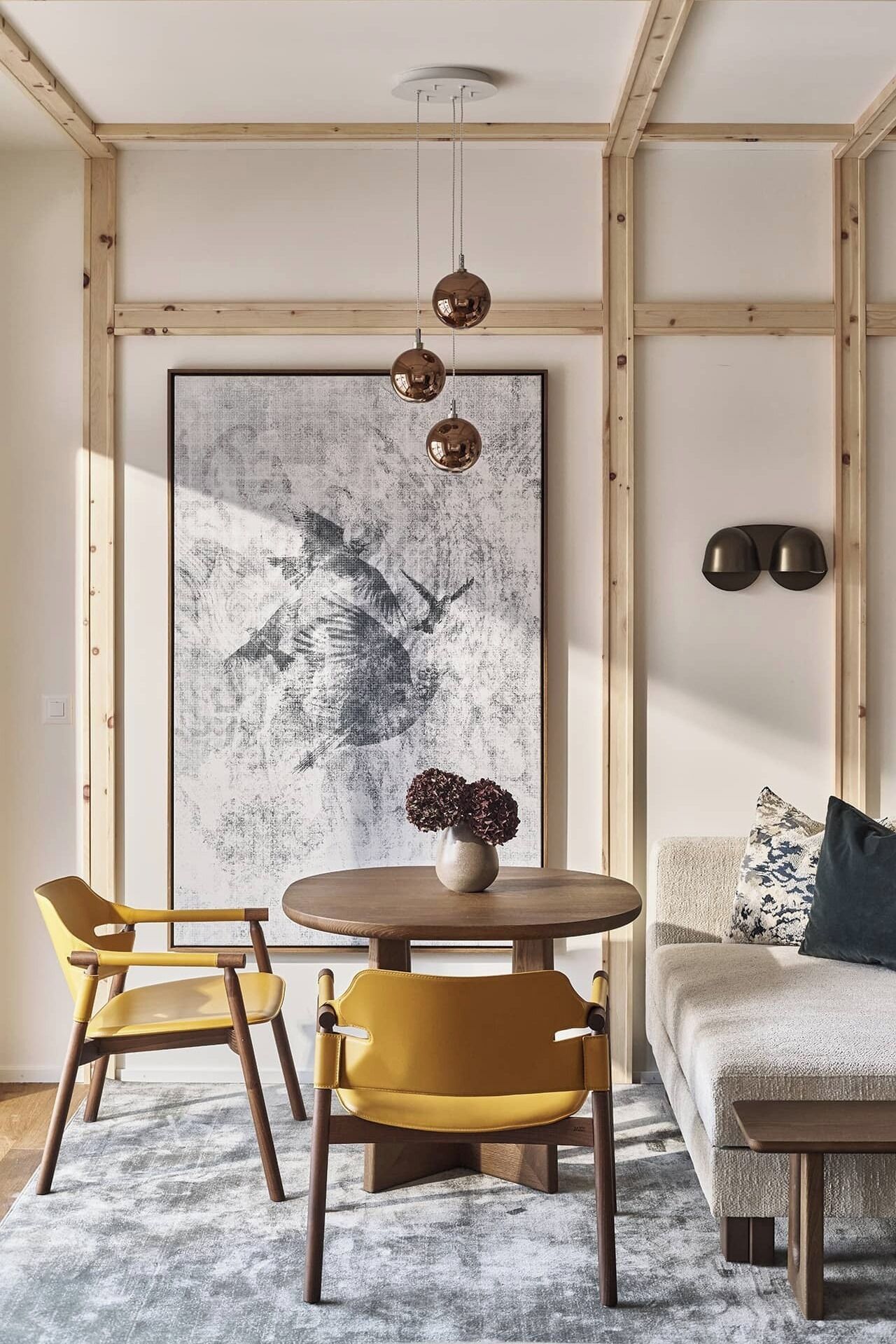 Ein modernes Wohnzimmer mit einem runden Holztisch, zwei gelben Stühlen, einem großen abstrakten Gemälde, einer Vase mit getrockneten Blumen und einer Couch mit gemusterten Kissen.