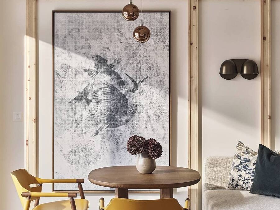 Ein modernes Wohnzimmer mit einem runden Holztisch, zwei gelben Stühlen, einem großen abstrakten Gemälde, einer Vase mit getrockneten Blumen und einer Couch mit gemusterten Kissen.