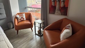 Uma sala de estar moderna com duas poltronas de couro, uma mesinha lateral e uma janela grande com vista para um edifício de telhado vermelho.