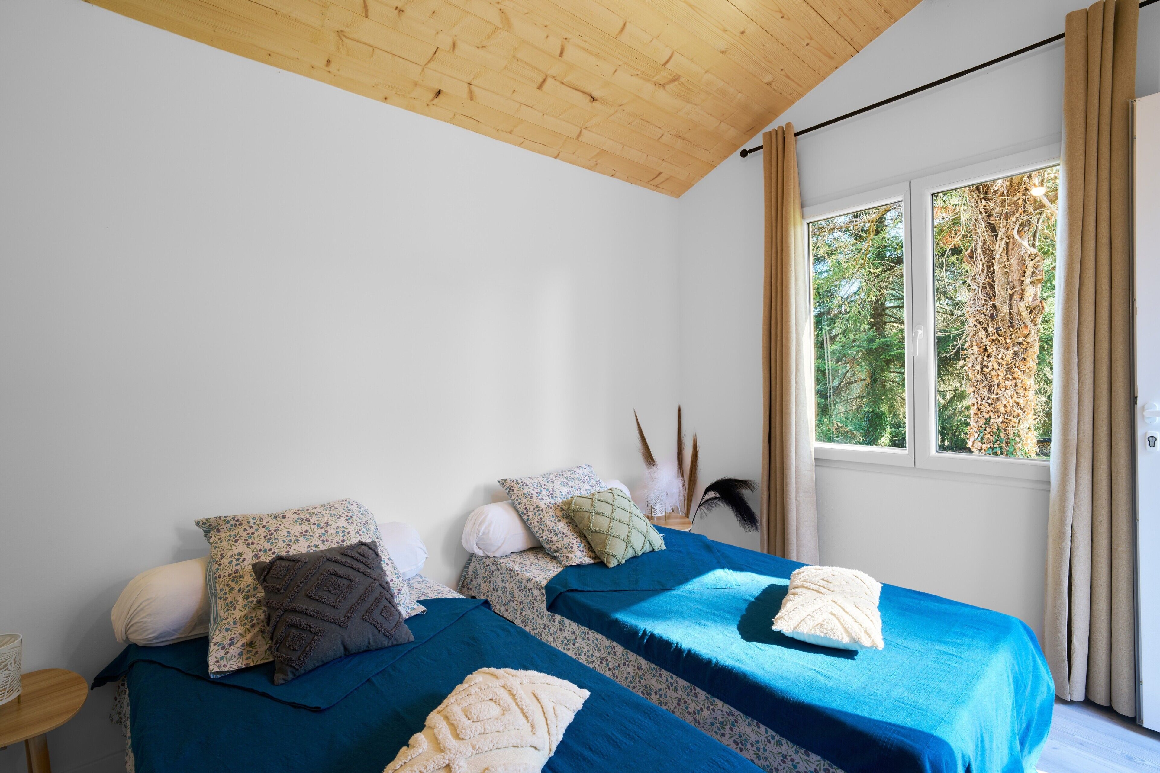 A room with two beds, a wooden ceiling, a window with curtains, and a view of trees.