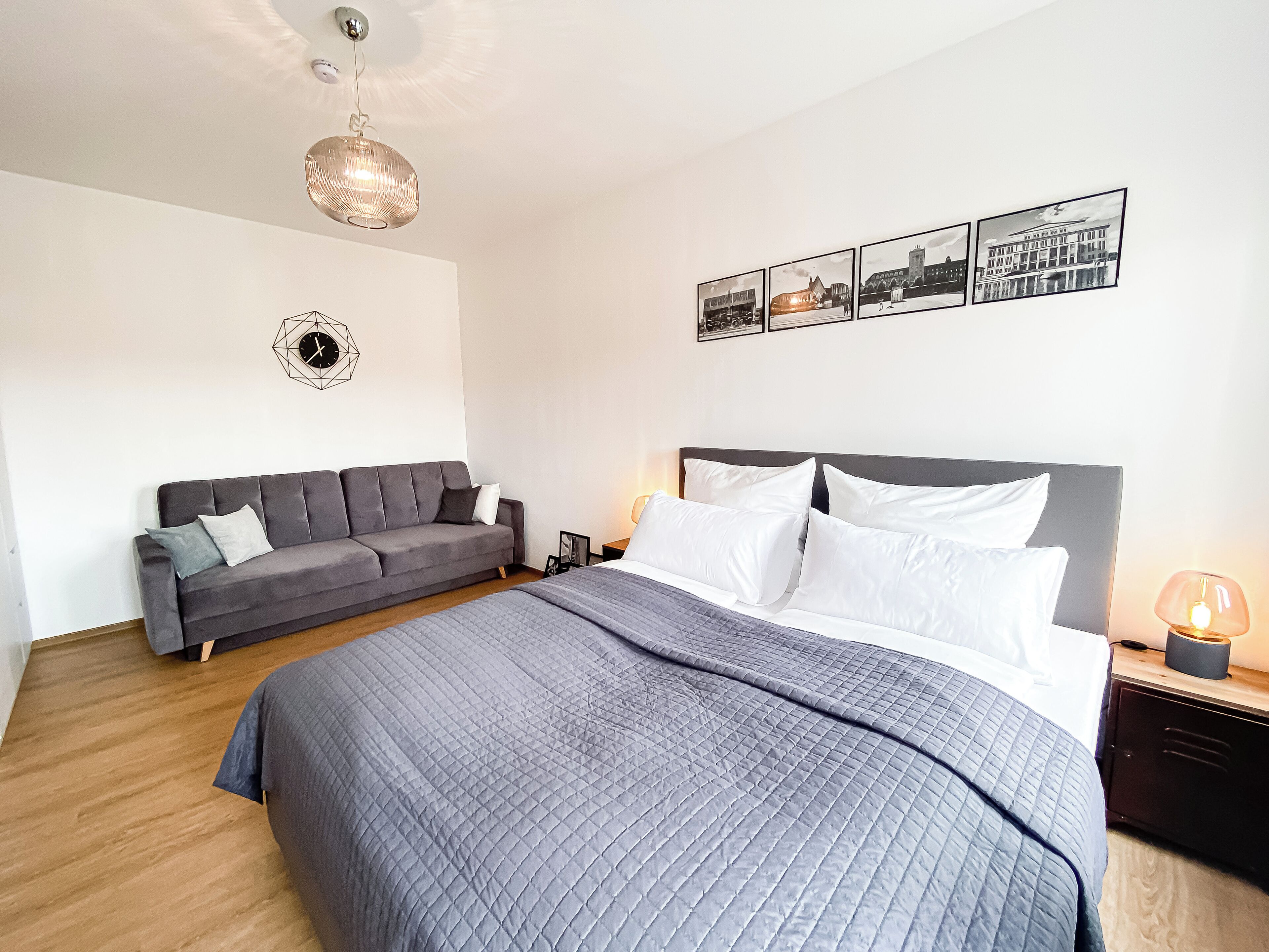 Une chambre moderne avec un grand lit, un canapé gris, une horloge murale et des tableaux encadrés.