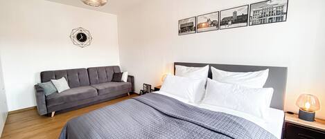 Une chambre moderne avec un grand lit, un canapé gris, une horloge murale et des tableaux encadrés.