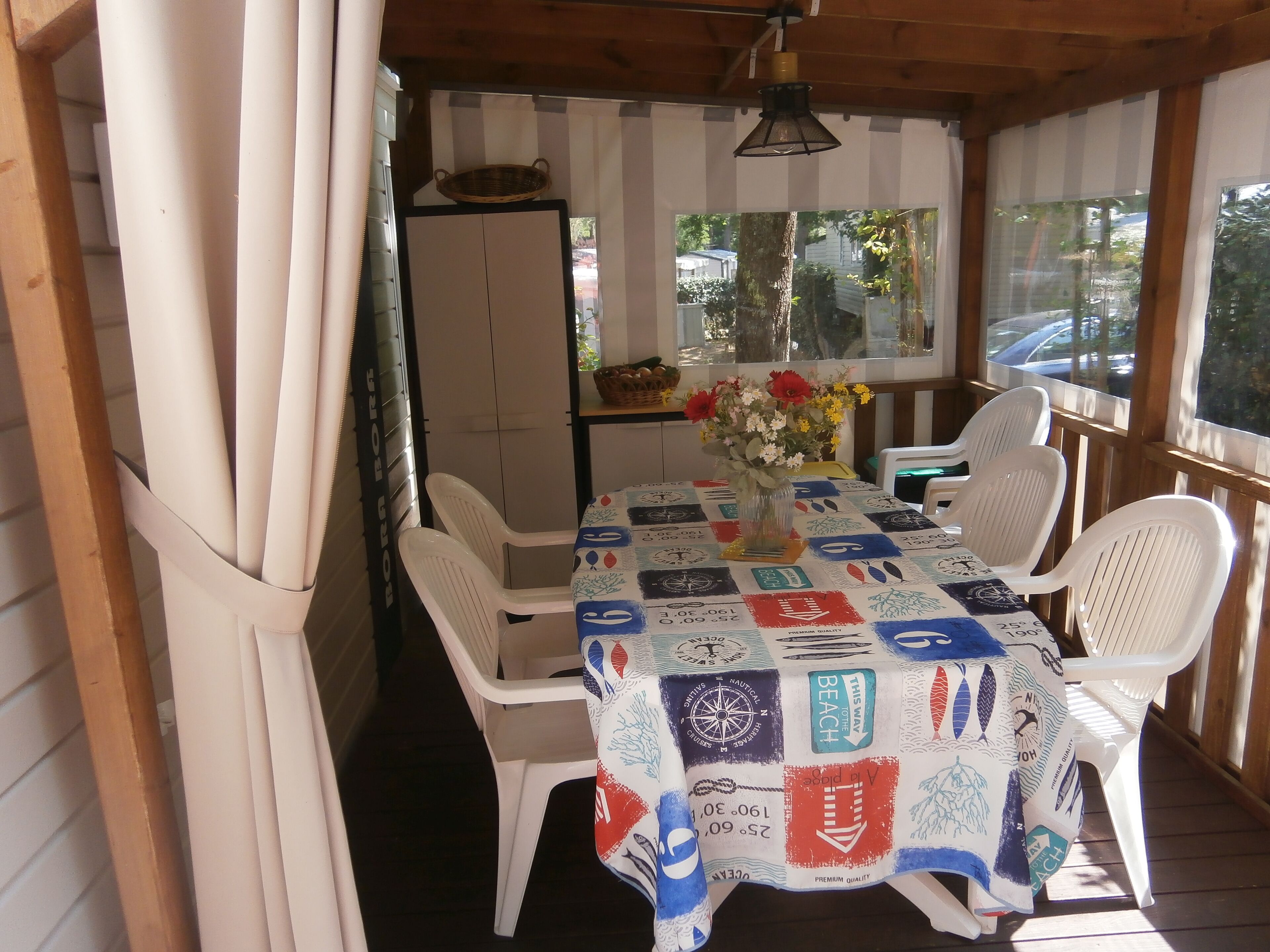 A covered outdoor dining area with a table set for four, surrounded by white plastic chairs. A vase with flowers is on the table, and a basket is on the wall behind the table.