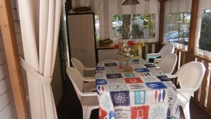 A covered outdoor dining area with a table set for four, surrounded by white plastic chairs. A vase with flowers is on the table, and a basket is on the wall behind the table. - Comfortable 3-bedroom mobile home near the beach (Saint-Brevin-les-Pins)
