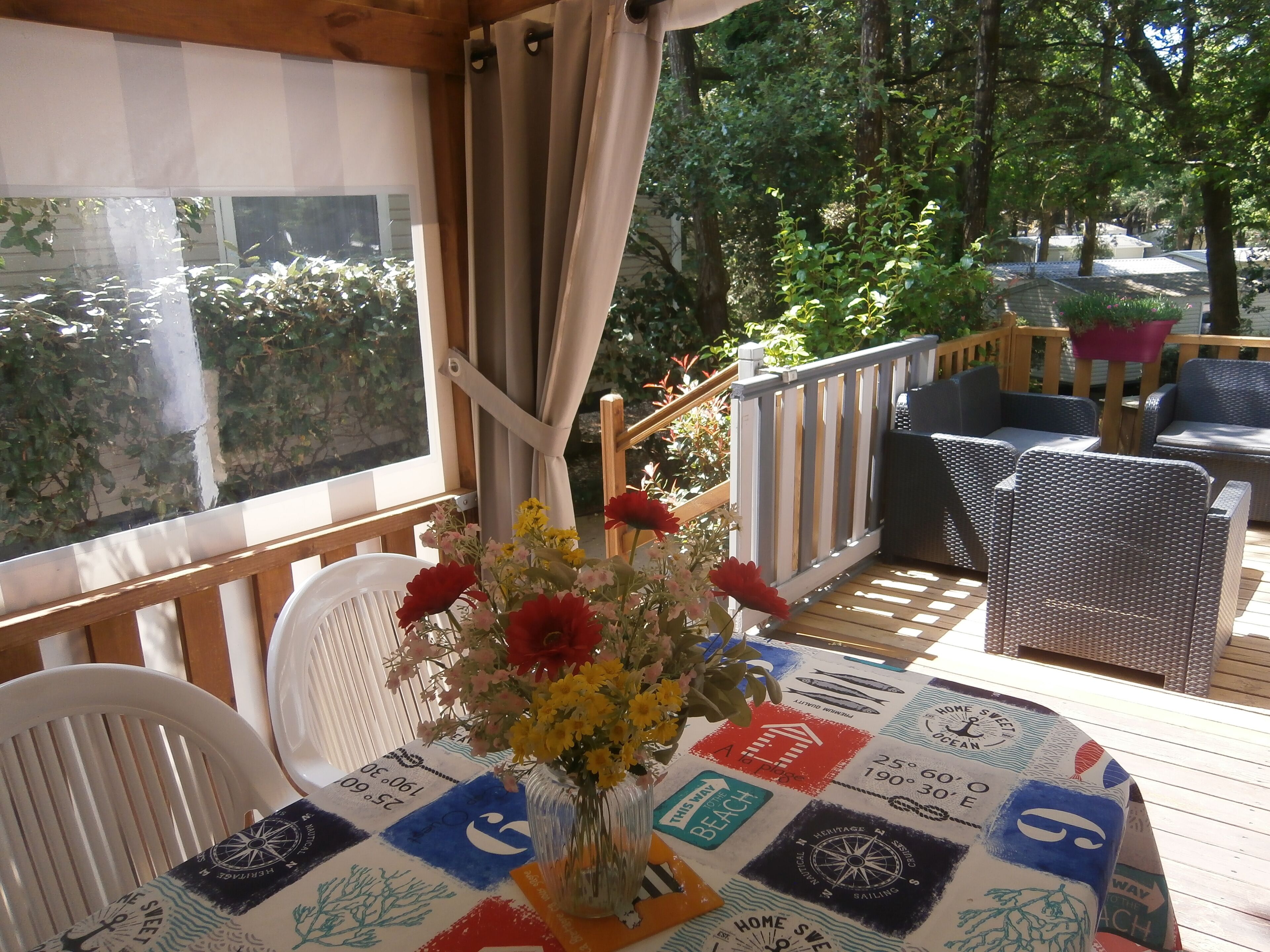 A covered outdoor seating area with a table set for two, featuring a nautical-themed tablecloth and a vase of flowers.