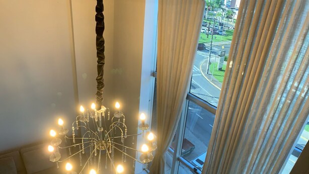 Een kroonluchter met meerdere lampen die aan een centraal punt hangt, een trap met metalen leuningen, en een uitzicht op een straat door een groot raam met gordijnen.