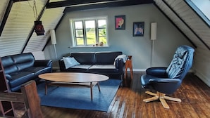 A loft-style room with a wooden floor, a sofa, a coffee table, and a blue armchair. There is a window with a view of greenery outside. - Nature, tranquillity and petting zoo (Vissenbjerg)