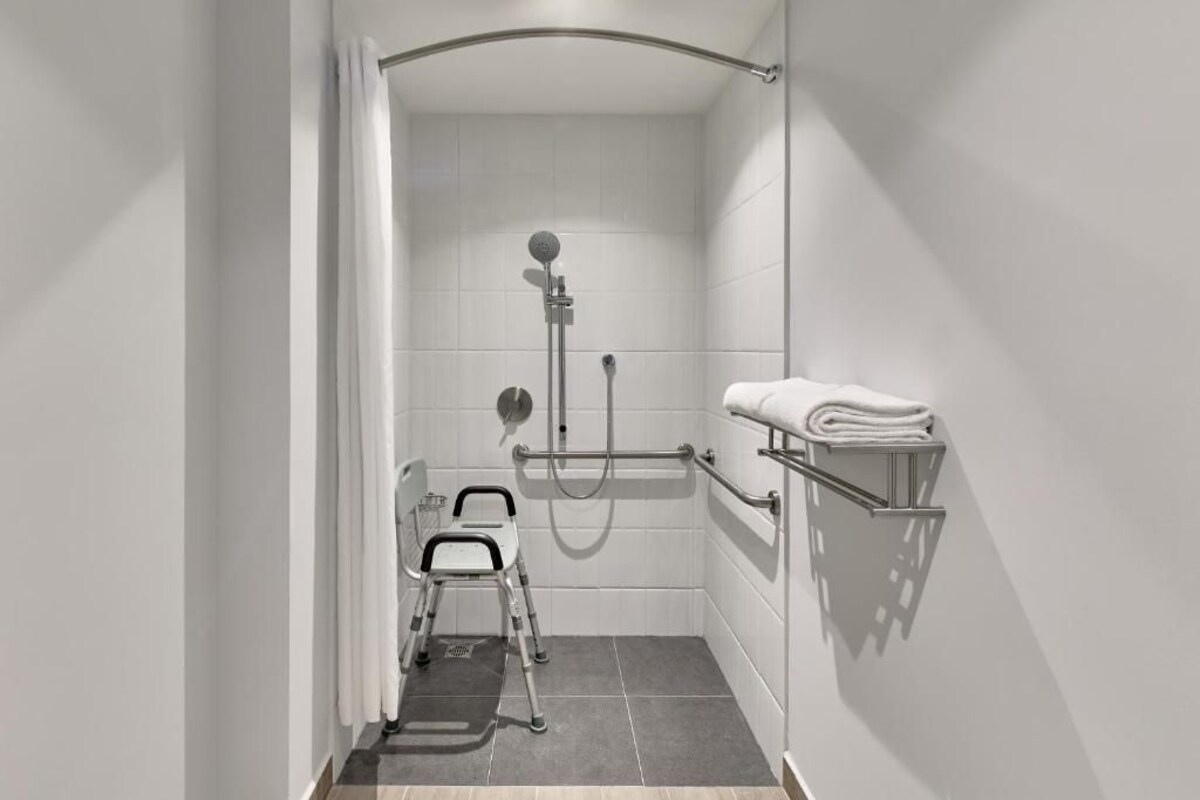 Een moderne badkamer met witte tegels, een douche met handdouche, een handgreep, een rolstoelvriendelijke stoel en een handdoekrek met opgevouwen handdoeken.