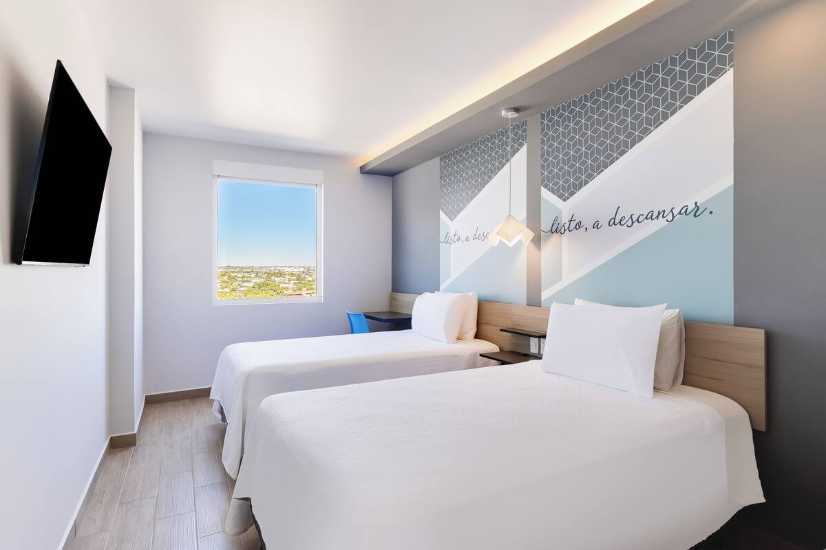 Ein Hotelzimmer mit zwei Betten, einem Fernseher, einem Fenster mit Blick und einer Wand mit geometrischem Muster und Text.