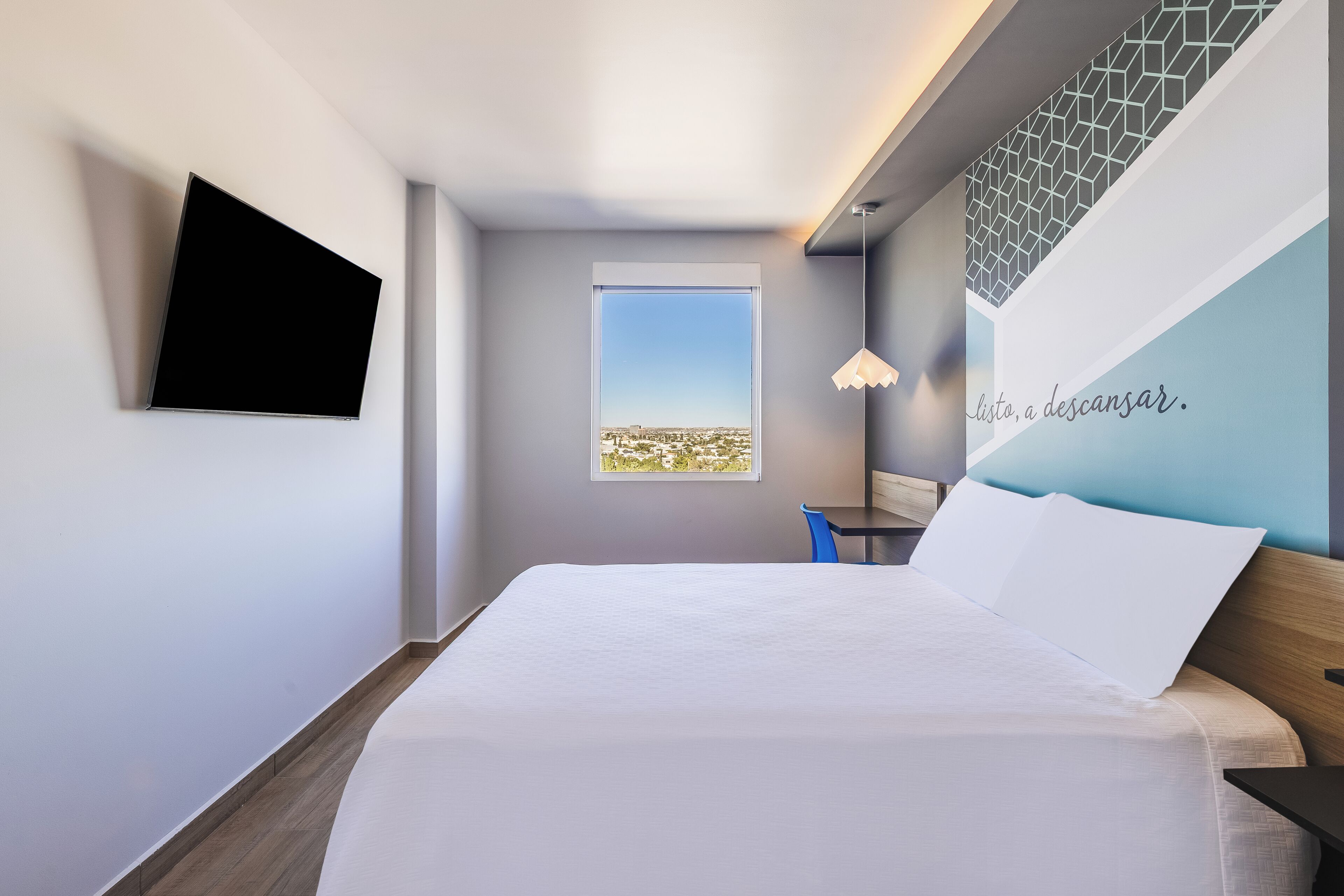 Ein modernes Hotelzimmer mit Bett, Wandfernseher, kleinem Tisch mit Stuhl, Fenster mit Stadtblick und dekorativer Wand mit Text.