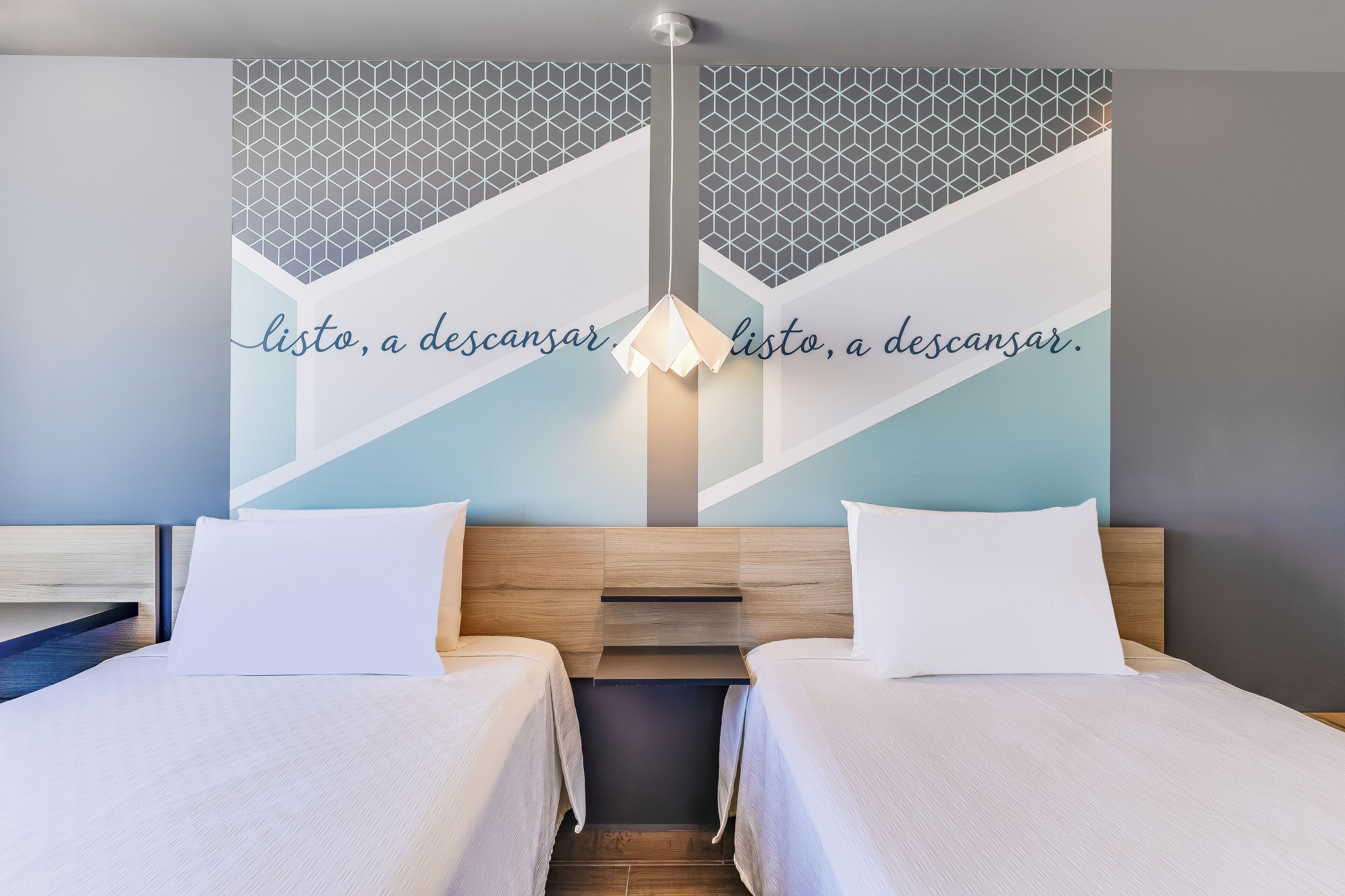 Une chambre d’hôtel avec deux lits, un motif géométrique sur le mur et un luminaire.