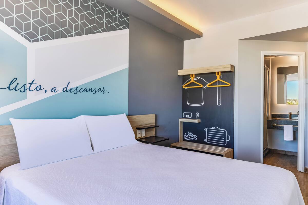 Ein Hotelzimmer mit einem Bett, einem an der Wand befestigten Kleiderhaken und einem durch eine geöffnete Tür einsehbaren Badezimmer.