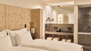A modern hotel room with a wooden headboard, a large bed, a bathroom with a sink and mirror, and a shower area. - Laudinella Apartments (St. Moritz)