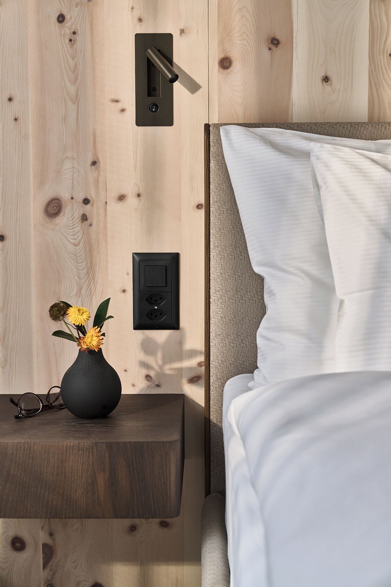 A wooden wall with a power outlet and a switch, a bedside table with a vase of flowers, and a bed with white bedding.