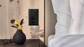 A wooden wall with a power outlet and a switch, a bedside table with a vase of flowers, and a bed with white bedding.