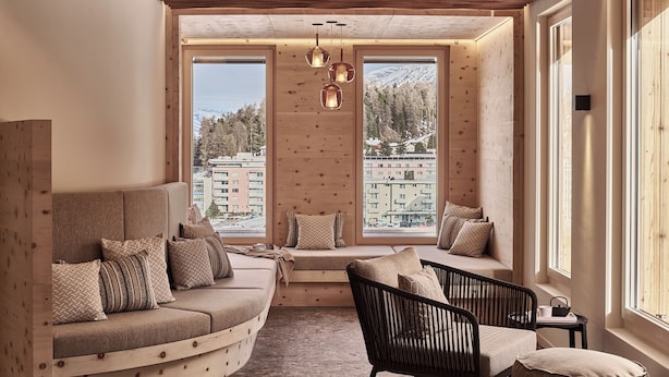 A cozy room with a curved sofa, a black chair, and a small table. Large windows offer a view of buildings and trees. - Laudinella Apartments (St. Moritz)