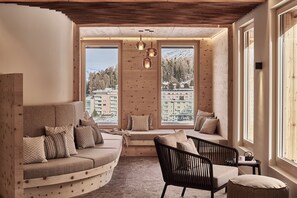 A cozy room with a curved sofa, a black chair, and a small table. Large windows offer a view of buildings and trees. - Laudinella Apartments (St. Moritz)