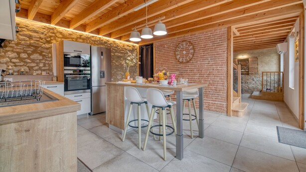 A modern kitchen with a stone wall, wooden beams, and a central island with bar stools. - Holiday Home 'Les Echaunes' with Private Terrace, Wi-Fi and Air Conditioning (Caraman)